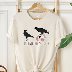 以下が含まれることがあります： 2 羽の黒いカラスが描かれたクリーム色のTシャツ。1 羽は立っており、もう 1 羽はピンクの花に囲まれた頭蓋骨の上に止まっています。「ATTEMPTED MURDER」の文字が下に印刷されています。木製のハンガーにTシャツが掛けられています。