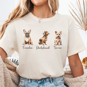 May include: A beige t-shirt with a watercolor illustration of three dog breeds: a French Bulldog, a Dachshund, and a Yorkshire Terrier. The text "Frenchie", "Dachshund", and "Terrier" is printed below each dog.