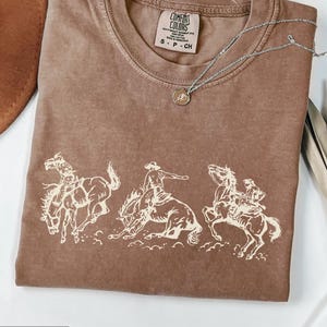 Puede incluir: Camiseta color espresso con cuello redondo y un gr&aacute;fico de vaqueros montando caballos salvajes. La camiseta tiene un aspecto vintage. Un collar plateado con un peque&ntilde;o colgante est&aacute; sobre la camiseta.
