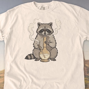 May include: Ivory-colored t-shirt featuring a cartoon raccoon smoking a bong. The raccoon is depicted in shades of gray and brown, with smoke rising from the bong. The shirt has a classic crew neck and short sleeves.