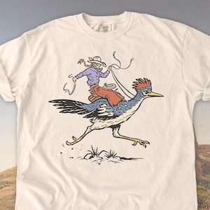May include: Ivory-colored t-shirt featuring a cartoon illustration of a person riding a roadrunner. The rider wears a purple shirt, red skirt, and a cowboy hat, holding a rope. The roadrunner is blue, white, and black, with a red crest. The shirt has a classic crew neck.
