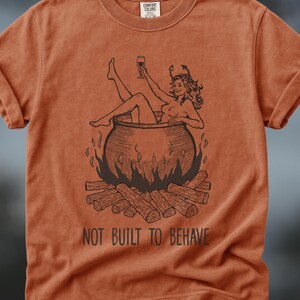 May include: A rust-colored short-sleeve t-shirt featuring a black line-art graphic of a figure with horns sitting in a cauldron over a fire while holding a wine glass. The text NOT BUILT TO BEHAVE is printed in black below the graphic illustration.