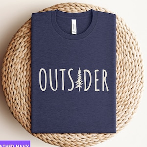 May include: Heather navy t-shirt with the word "OUTSIDER" in white, featuring a tree graphic. The shirt is folded and placed on a woven, circular surface. The word "CANVAS" is printed on the tag.
