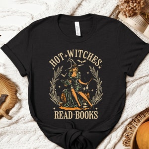 May include: Black t-shirt with a vintage-style graphic featuring a witch in a hat, sitting on a pumpkin, with the text "HOT WITCHES READ BOOKS". The design includes stars, bats, and a decorative border.
