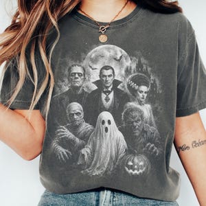 May include: A charcoal gray short-sleeve t-shirt featuring a detailed monochromatic graphic print of classic horror movie monsters, including a vampire, a mummy, a ghost, a werewolf, and a pumpkin, set against a full moon and castle background.