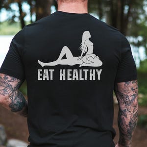 May include: Black t-shirt with a white graphic of a reclining figure and the text "EAT HEALTHY". The design features a silhouette of a person in a relaxed pose. The shirt is made of a soft material.