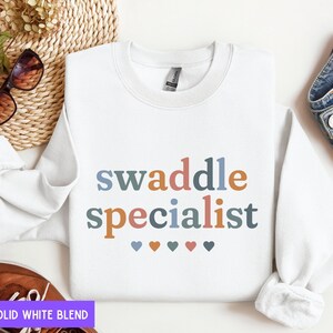 May include: White sweatshirt with the text "swaddle specialist" in colorful letters and five small hearts below. The text is in a playful, casual font.