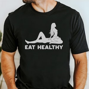 May include: Black t-shirt with a white graphic of a reclining figure and the text "EAT HEALTHY". The design features a stylized illustration of a person. The shirt is a crew neck.