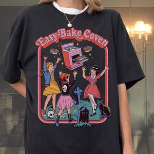 May include: Black t-shirt with the text "Easy-Bake Coven" and a retro graphic of three girls, a tombstone, and a black cat. The graphic features a pink oven and baked goods. The shirt is a relaxed fit.