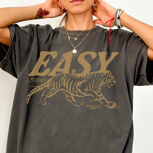 May include: Dark grey t-shirt with the word "EASY" in gold lettering above a gold tiger graphic. The shirt has a relaxed fit and short sleeves. The word "PEPPER" is at the bottom left.