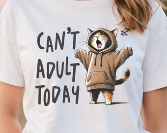 May include: White t-shirt with the phrase "CAN'T ADULT TODAY" in black lettering. The shirt features a cartoon cat wearing a brown hoodie with its arms outstretched. The cat has orange, black, and white fur.