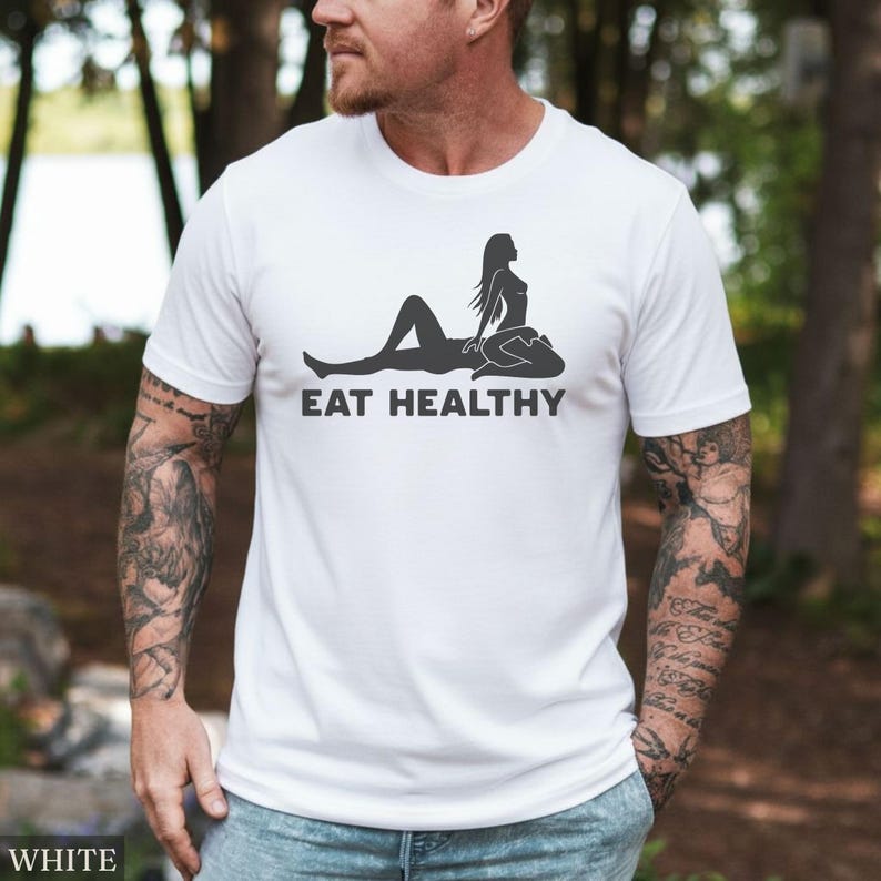 May include: White t-shirt with a black graphic of a reclining figure and the text "EAT HEALTHY". The shirt is made of a soft material and has short sleeves. The word "WHITE" is visible at the bottom left.
