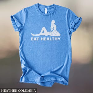 May include: A light blue t-shirt with the words "EAT HEALTHY" in white, along with a white graphic of a person. The shirt is made of a soft material and has rolled-up sleeves. The text "HEATHER COLUMBIA" is at the bottom.