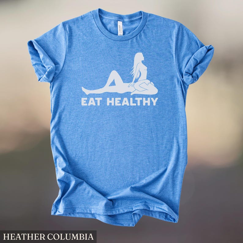 May include: A light blue t-shirt with the words "EAT HEALTHY" in white, along with a white graphic of a person. The shirt is made of a soft material and has rolled-up sleeves. The text "HEATHER COLUMBIA" is at the bottom.