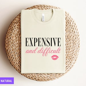 May include: A beige t-shirt with the text "EXPENSIVE and difficult" in black lettering and a pink lipstick print below. The shirt is displayed on a woven straw surface.