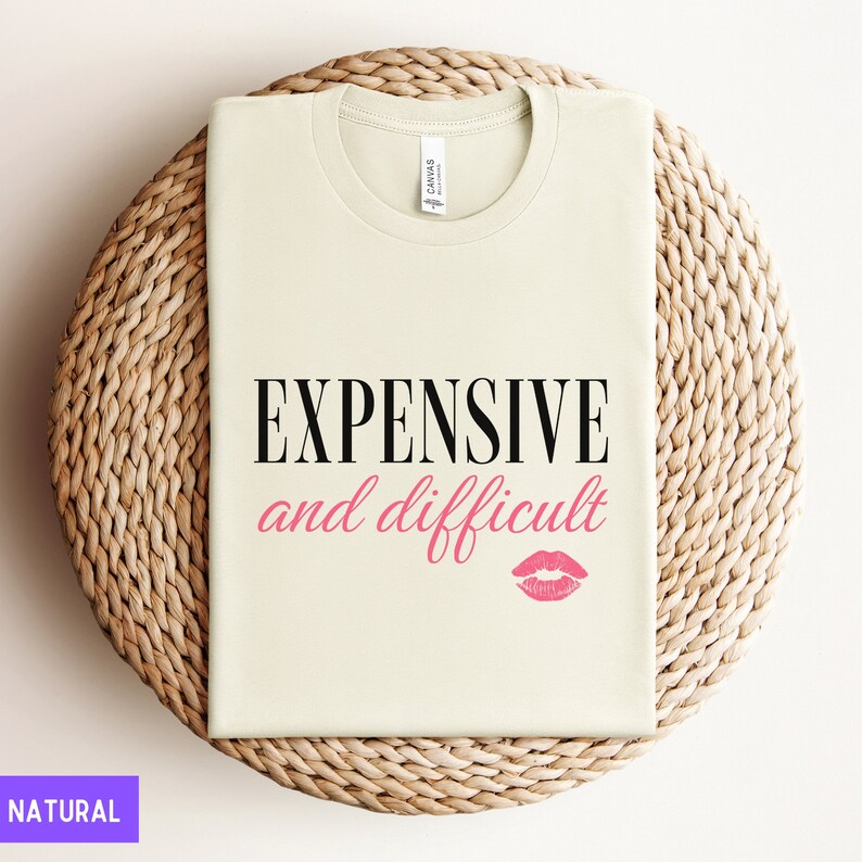 May include: A beige t-shirt with the text "EXPENSIVE and difficult" in black lettering and a pink lipstick print below. The shirt is displayed on a woven straw surface.