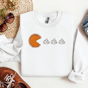 Pode incluir: Camisola branca com um gr&aacute;fico de uma fatia de torta de ab&oacute;bora comendo tr&ecirc;s colheres de sopa de chantilly. O gr&aacute;fico est&aacute; no estilo do videogame Pac-Man.