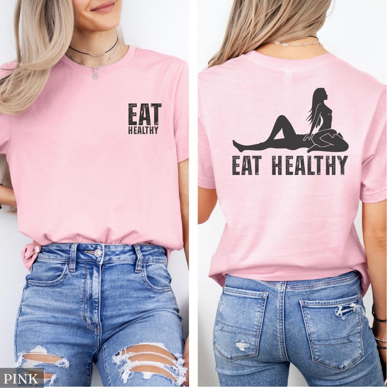 May include: Pink t-shirt with the text "EAT HEALTHY" on the front and back. The back of the shirt features a silhouette of a person in a yoga pose above the text. The shirt is paired with blue jeans.