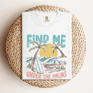 May include: White t-shirt with a graphic design of palm trees, waves, and a surfboard. The text "FIND ME UNDER THE PALMS" is printed on the shirt.