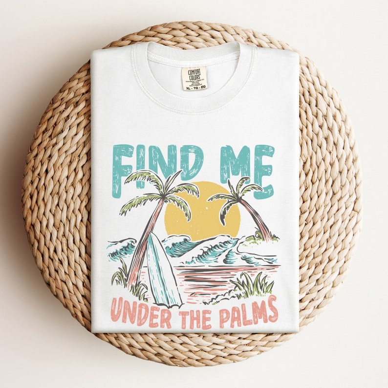 May include: White t-shirt with a graphic design of palm trees, waves, and a surfboard. The text "FIND ME UNDER THE PALMS" is printed on the shirt.