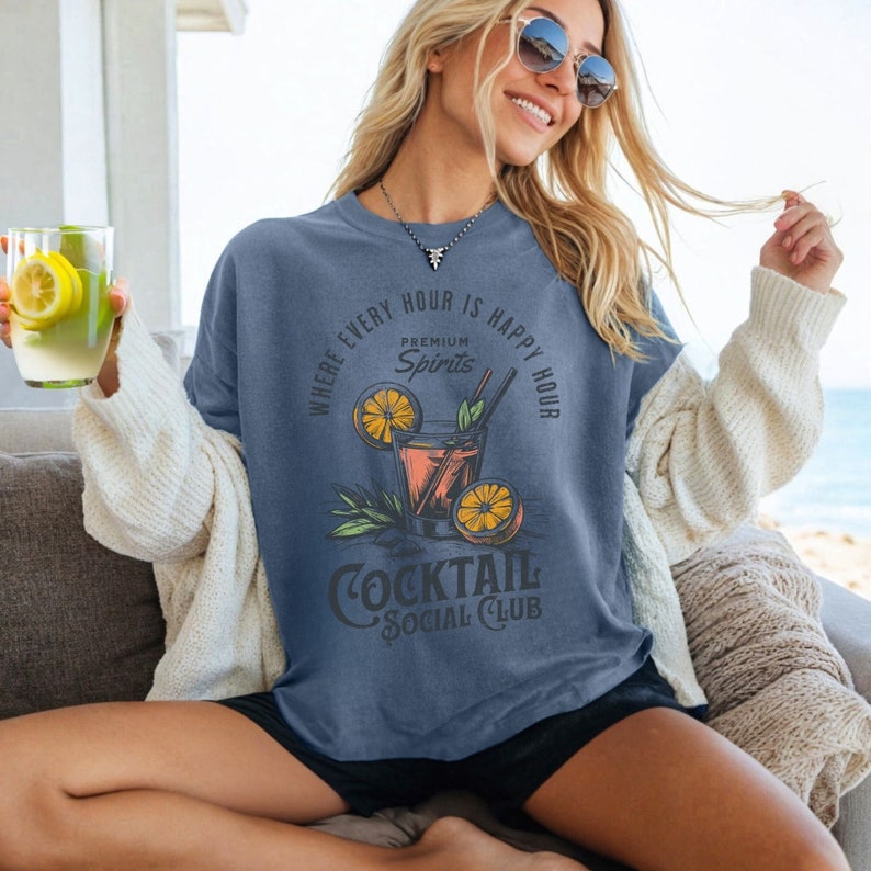May include: A blue t-shirt with a graphic of a cocktail glass with orange slices and the text "Where every hour is happy hour, Premium Spirits, Cocktail Social Club".