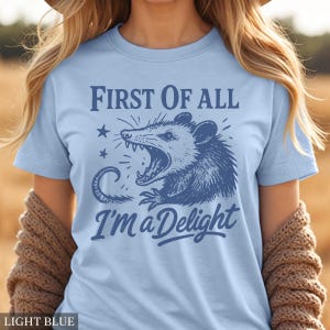May include: Light blue t-shirt with a distressed blue graphic of an opossum with the text "First of All I'm a Delight". The opossum is depicted with its mouth open, surrounded by stars and a curved line.
