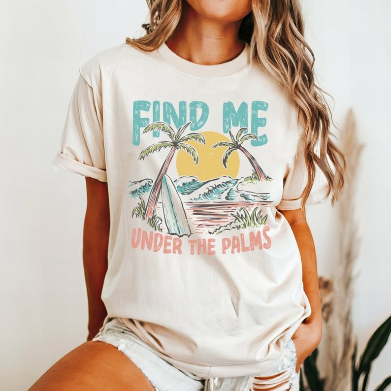May include: A beige t-shirt with a graphic print of palm trees, a surfboard, and waves. The text "FIND ME UNDER THE PALMS" is printed in blue, pink, and yellow.