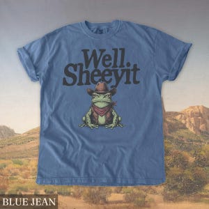 May include: Blue jean t-shirt featuring a cartoon frog wearing a cowboy hat, vest, and bandana. The text "Well. Sheeeyit" is printed above the frog. The shirt has rolled-up sleeves and the words "BLUE JEAN" are printed at the bottom.