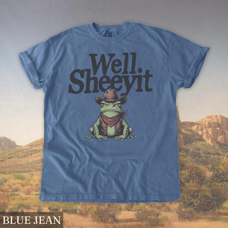 May include: Blue jean t-shirt featuring a cartoon frog wearing a cowboy hat, vest, and bandana. The text "Well. Sheeeyit" is printed above the frog. The shirt has rolled-up sleeves and the words "BLUE JEAN" are printed at the bottom.