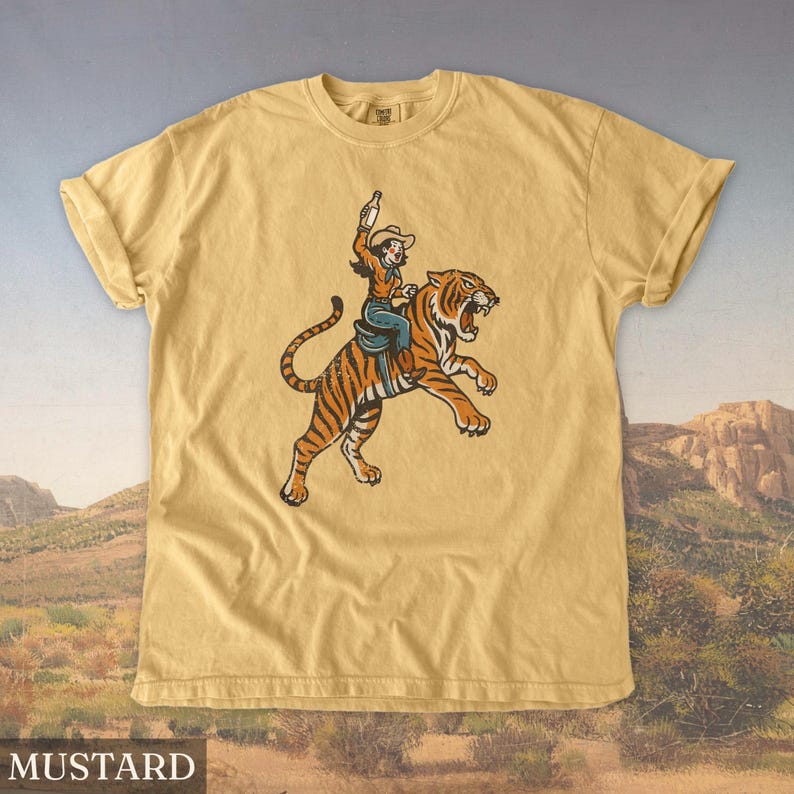 May include: Mustard-colored t-shirt featuring a graphic of a person riding a tiger, holding a bottle. The tiger is orange and black, and the rider is wearing a cowboy hat and blue jeans. The shirt has a classic crew neck and short sleeves.