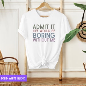 May include: White t-shirt with the text "ADMIT IT LIFE WOULD BE BORING WITHOUT ME" in green, blue, and purple. The shirt is hanging on a wooden hanger against a light background. The shirt is a solid white blend.