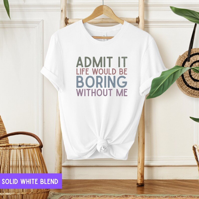 May include: White t-shirt with the text "ADMIT IT LIFE WOULD BE BORING WITHOUT ME" in green, blue, and purple. The shirt is hanging on a wooden hanger against a light background. The shirt is a solid white blend.
