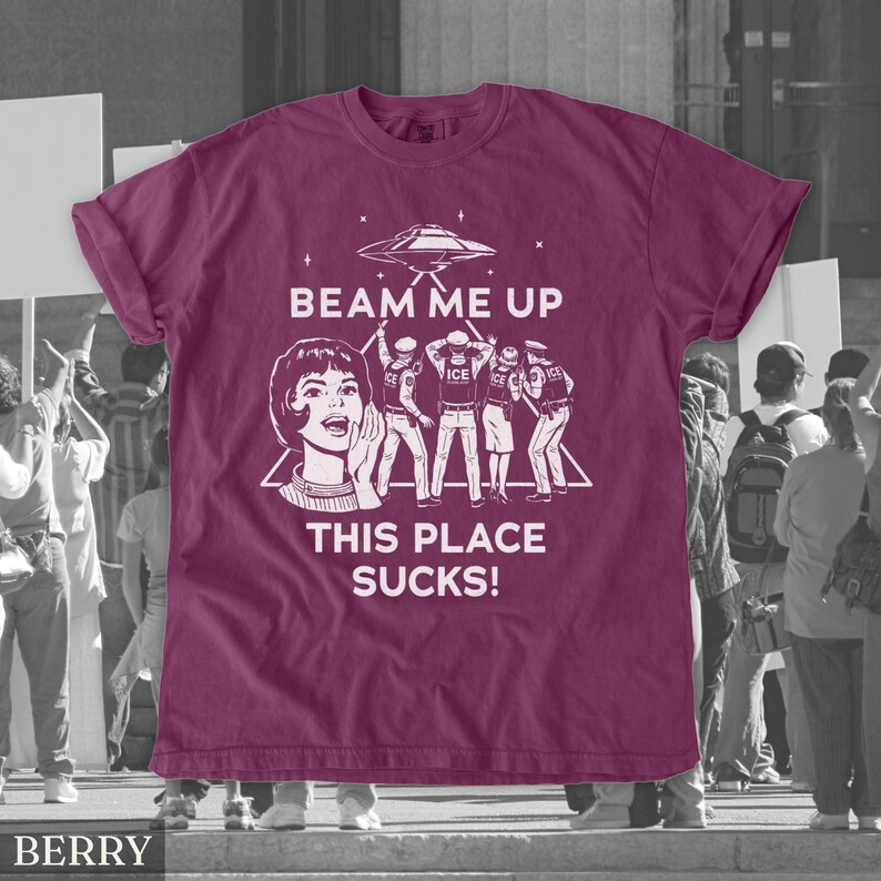 May include: Berry-coloured t-shirt with a white graphic design. The design includes a retro illustration of a woman, an alien spaceship, and figures with the text "BEAM ME UP THIS PLACE SUCKS!"