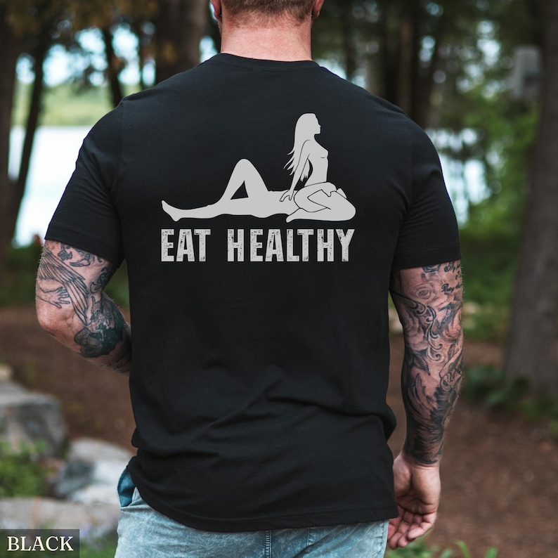 May include: Black t-shirt with a white graphic of a reclining figure and the text "EAT HEALTHY". The design features a silhouette of a person in a relaxed pose. The shirt is made of a soft material.