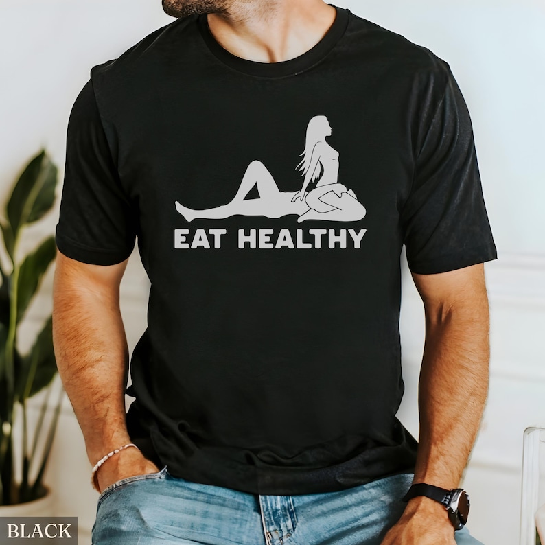 May include: Black t-shirt with a white graphic of a reclining figure and the text "EAT HEALTHY". The design features a stylized illustration of a person. The shirt is a crew neck.