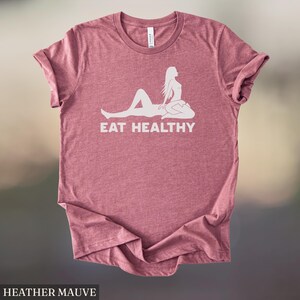 May include: Heather mauve t-shirt with a white graphic of a reclining figure and the words "EAT HEALTHY". The shirt is made of a soft material and has a classic crew neck design. The shirt is a solid color.