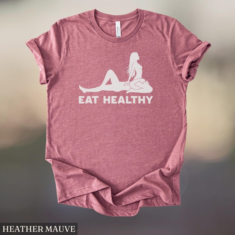 May include: Heather mauve t-shirt with a white graphic of a reclining figure and the words "EAT HEALTHY". The shirt is made of a soft material and has a classic crew neck design. The shirt is a solid color.