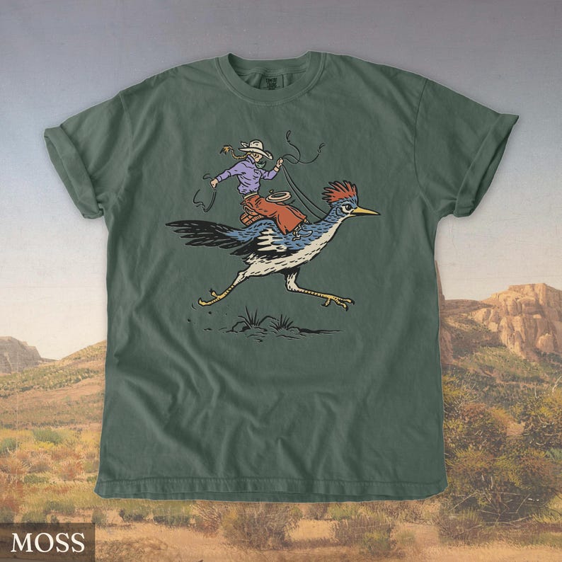 May include: Moss green t-shirt featuring a cartoon illustration of a cowboy riding a roadrunner. The cowboy wears a hat, purple shirt, and orange chaps, holding a lasso. The roadrunner is blue, white, and black. The word "MOSS" is in the bottom left corner.