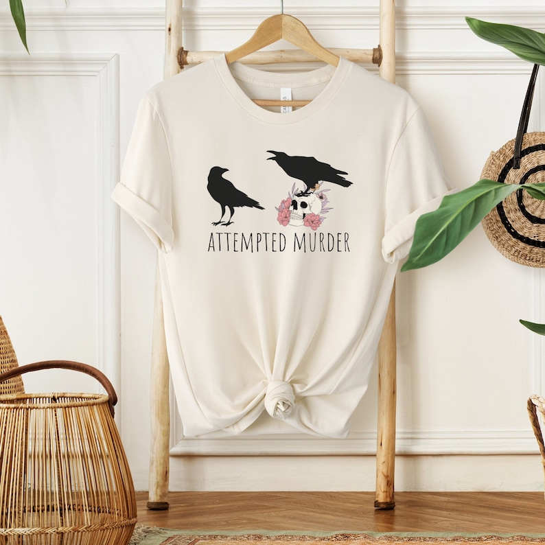 以下が含まれることがあります： 2 羽の黒いカラスが描かれたクリーム色のTシャツ。1 羽は立っており、もう 1 羽はピンクの花に囲まれた頭蓋骨の上に止まっています。「ATTEMPTED MURDER」の文字が下に印刷されています。木製のハンガーにTシャツが掛けられています。