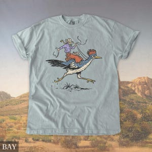 May include: Light blue t-shirt featuring a cartoon illustration of a cowboy riding a roadrunner. The cowboy wears a purple shirt, red pants, and a white hat. The roadrunner is blue, black, and white. The word "BAY" is at the bottom.