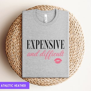 May include: A heather grey t-shirt with the text "EXPENSIVE and difficult" in black lettering and a pink lipstick print.