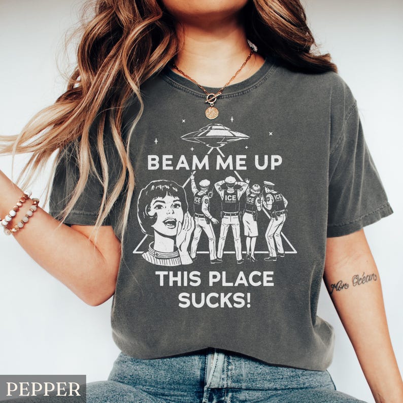 May include: Dark grey t-shirt with a graphic design featuring a retro woman, an alien spaceship, and the text "BEAM ME UP THIS PLACE SUCKS!". The design includes figures with the word "ICE" on their uniforms. The shirt is a casual wear item.