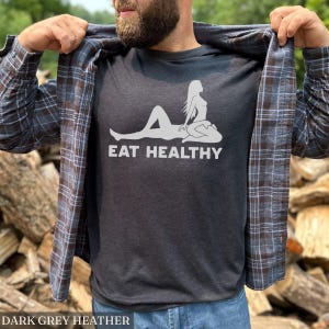May include: Dark grey heather t-shirt with a white graphic of a reclining figure and the words "EAT HEALTHY". The shirt is worn with a plaid button-down shirt and blue jeans. The text "DARK GREY HEATHER" is at the bottom of the image.