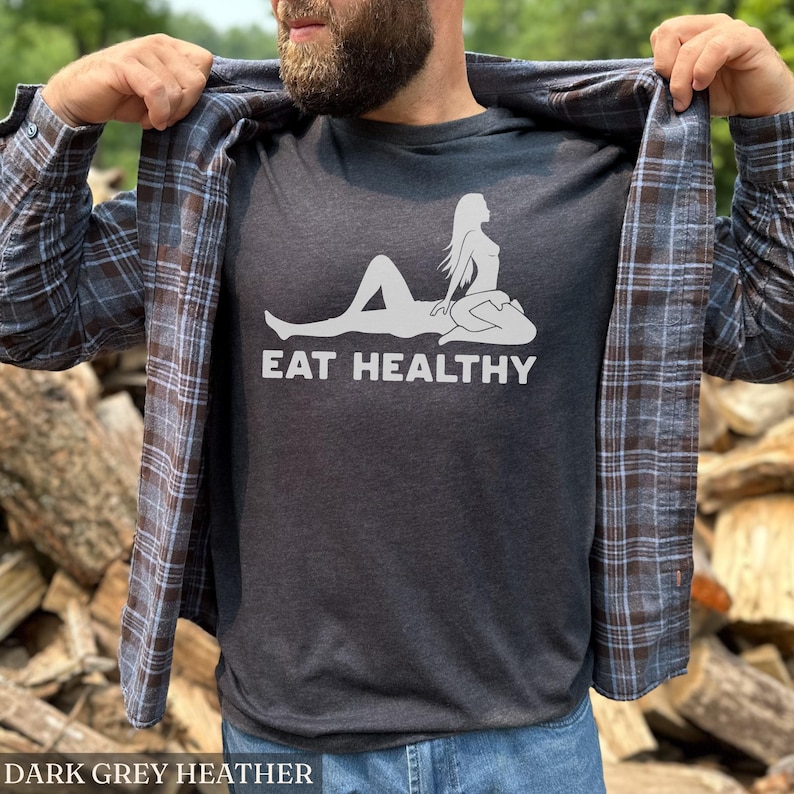 May include: Dark grey heather t-shirt with a white graphic of a reclining figure and the words "EAT HEALTHY". The shirt is worn with a plaid button-down shirt and blue jeans. The text "DARK GREY HEATHER" is at the bottom of the image.
