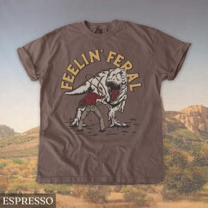 May include: Espresso-colored t-shirt featuring a graphic of a cowboy roping a Tyrannosaurus Rex. The text "FEELIN' FERAL" is arched above the dinosaur. The shirt is a short-sleeved crew neck.