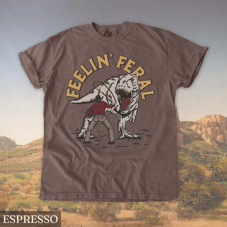 May include: Espresso-colored t-shirt featuring a graphic of a cowboy roping a Tyrannosaurus Rex. The text "FEELIN' FERAL" is arched above the dinosaur. The shirt is a short-sleeved crew neck.