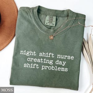 May include: A moss green t-shirt with the text "night shift nurse creating day shift problems" in white. The shirt has a crew neck and short sleeves. A gold necklace with a pendant is on the shirt.