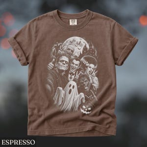 May include: A brown short-sleeve t-shirt featuring a white graphic print of classic horror movie monsters including Frankenstein, Dracula, a mummy, a werewolf, and a ghost, with a haunted castle and full moon in the background.