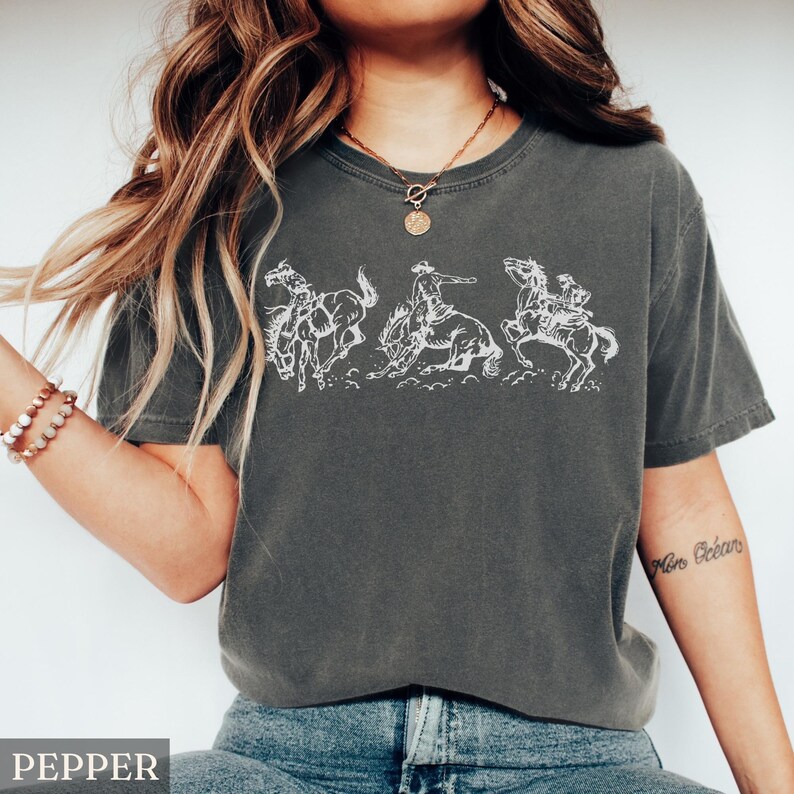 Puede incluir: Camiseta color pimienta con un gr&aacute;fico blanco de tres vaqueros montando caballos salvajes. El dise&ntilde;o es de estilo dibujo lineal. La camiseta tiene cuello redondo y mangas cortas. La palabra "PEPPER" est&aacute; impresa abajo a la izquierda.