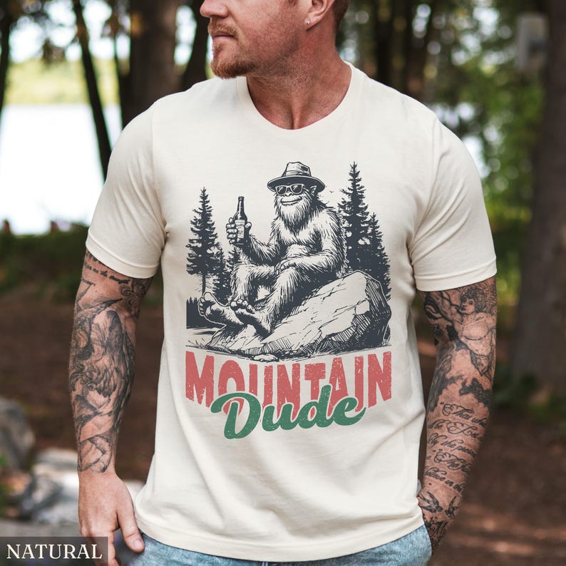 May include: Cream-colored t-shirt featuring a Bigfoot graphic wearing sunglasses and a hat, holding a beer bottle. The text "MOUNTAIN Dude" is printed in red and green. The shirt is made of a soft material.